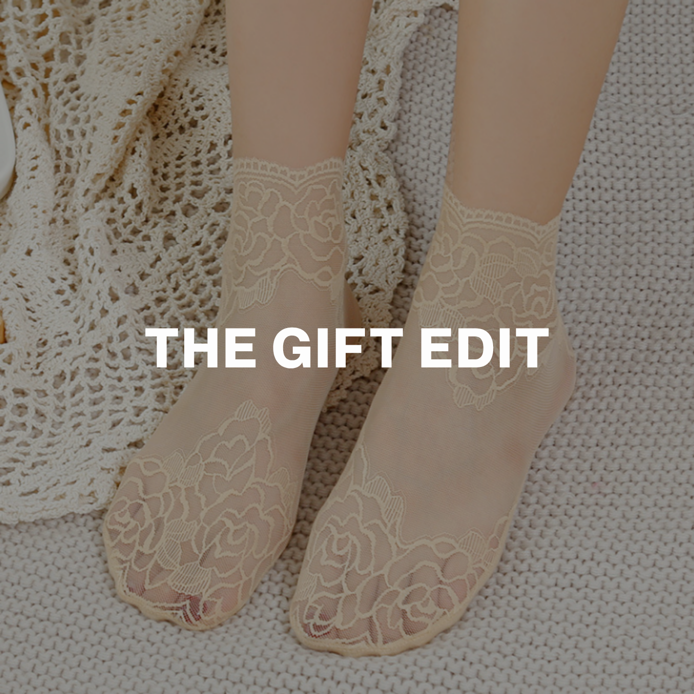 Model wearing sheer beige lace ankle socks with floral embroidery, styled on a knit blanket — labeled The Gift Edit.