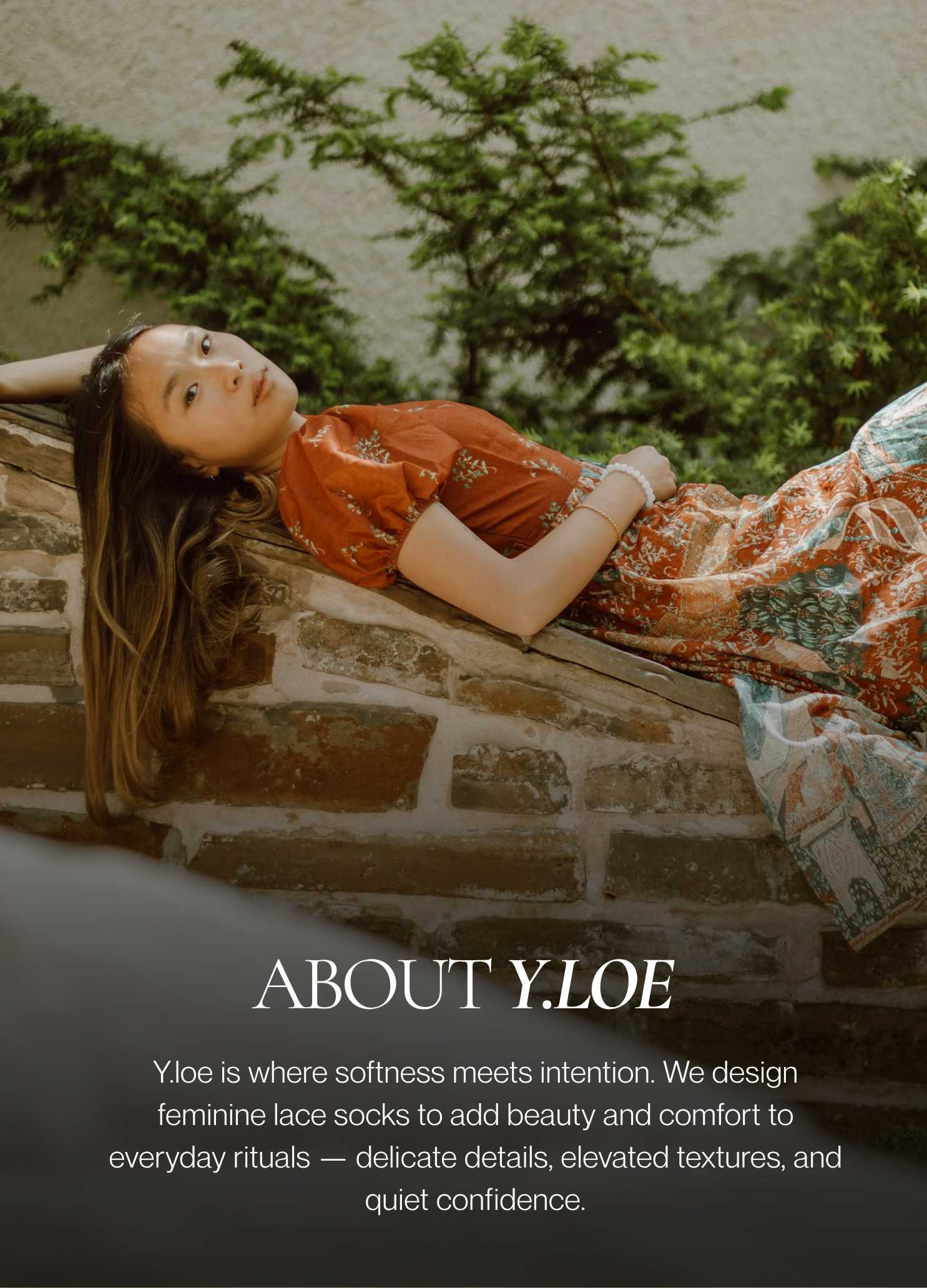 Woman lounging on a stone wall in a printed dress and white heels, surrounded by greenery — featured in the About Y.loe section.