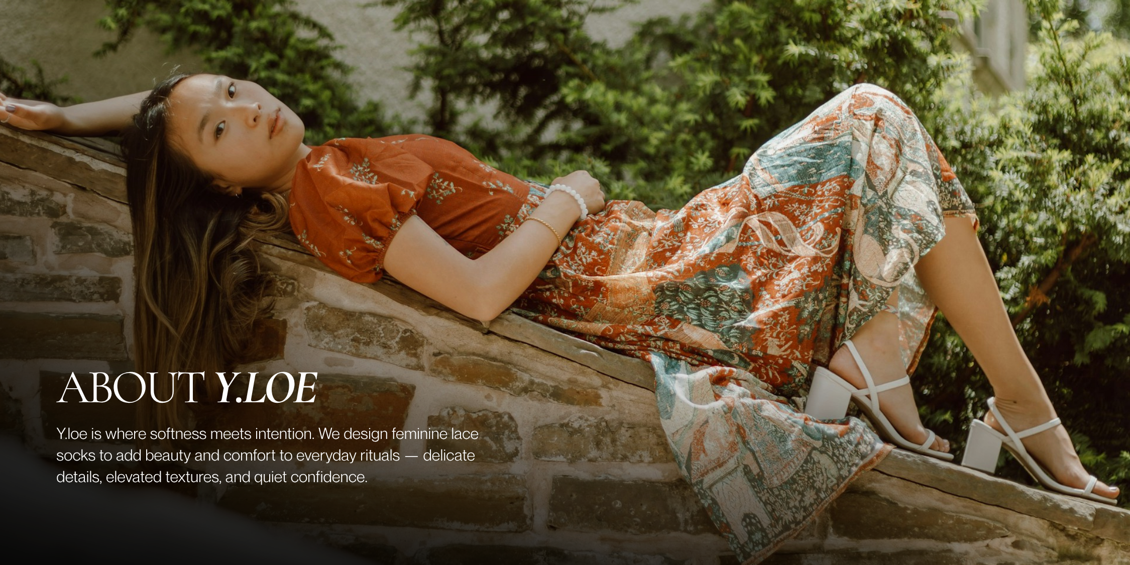 Woman lounging on a stone wall in a printed dress and white heels, surrounded by greenery — featured in the About Y.loe section.